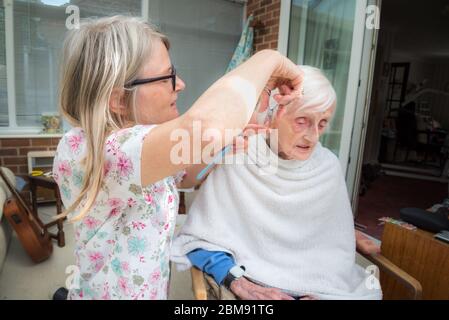 Covoid 19 restrizioni significa che la vecchia signora non può essere presa al parrucchiere come al solito, in modo che la sua caregiver visita rifinge e combatte i suoi capelli per lei come ge Foto Stock