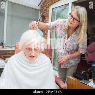 Covoid 19 restrizioni significa che la vecchia signora non può essere presa al parrucchiere come al solito, in modo che la sua caregiver visita rifinge e combatte i suoi capelli per lei come ge Foto Stock