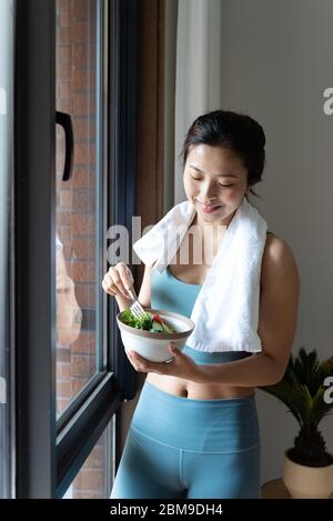 Una giovane donna asiatica che mangia verdure verdi sane vicino alla finestra Foto Stock