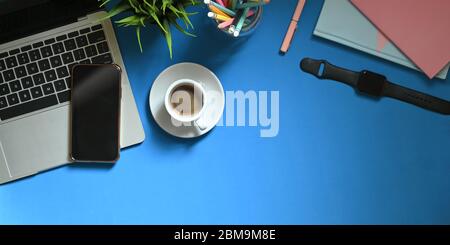 Immagine dall'alto del computer portatile che si mette sulla scrivania colorata con tazza da caffè, piante in vaso, matite in vaso di vetro, smartphone, nota, smartwatch Foto Stock