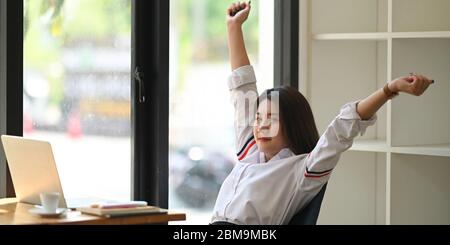 Foto di una bella donna che lavora come segretaria alzò le mani e la stretching mentre si siede e si rilassava al tavolo da lavoro in legno su comodo Foto Stock