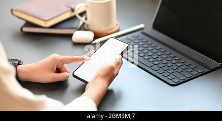 Immagine ritagliata delle mani di una donna elegante che tiene uno smartphone bianco sullo schermo vuoto sopra il tablet del computer che mette sulla scrivania circondato da caffè Foto Stock