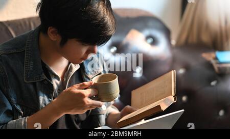 Uomo di primo piano che legge un libro e tiene una tazza di caffè in mano mentre siede al divano di pelle sopra il soggiorno comodo come sfondo. Un uomo ha ottenuto Foto Stock