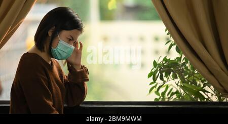 Foto di giovane donna che indossa la maschera medica sensazione di mal di testa mentre si trova alle finestre del moderno soggiorno. Donna che tiene una quarantena lei stessa a. Foto Stock