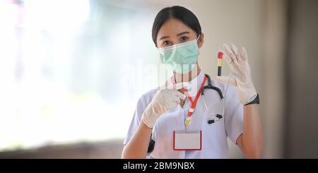 Medico femminile che indossa una maschera medica e un guanto che mostra un tubo di sangue per l'esame mentre si trova nella moderna sala d'esame sopra l'ospedale offuscato come ba Foto Stock