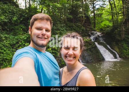 coppia sorridente che prende selfie davanti alla cascata nei boschi Foto Stock