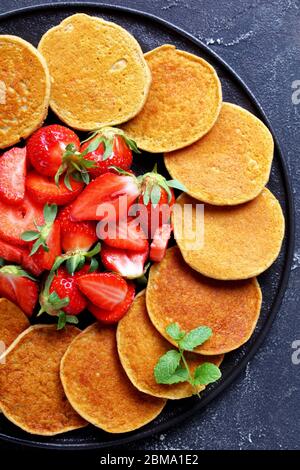 pancake senza glutine d'avena con fragole e miele su un piatto nero su un tavolo di cemento, cibo sano, vista verticale dall'alto Foto Stock