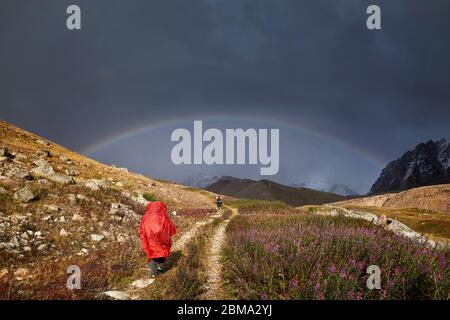 Turista nella Red Rain cover è a piedi giù per la strada di montagna di arcobaleno a sfondo Foto Stock