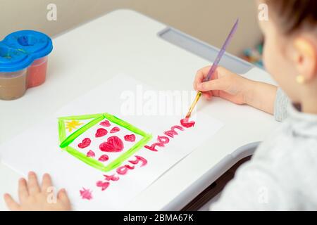 Bambino sta disegnando le parole soggiorno a casa sotto casa dipinta con i cuori rossi da acquerelli su foglio bianco di carta. La creatività dei bambini in quarantena. Foto Stock