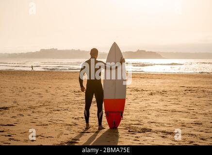 Vista posteriore del surfista atleta con surf sulla spiaggia al tramonto o all'alba. Silhouette di uomo surf guardando l'oceano in attesa di onde alte. Estrema Foto Stock