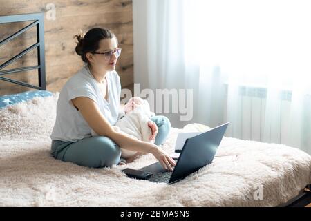 Lavoro a distanza, allenamento, lavoro a casa, femminilità, tecnologia, tempo libero, comunicazione concetto online - giovane ragazza carina con neonato si siede a letto Foto Stock