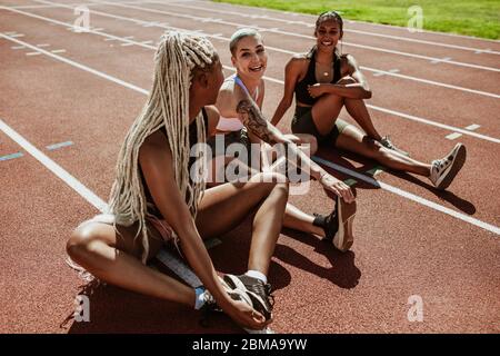 Gruppo felice di atleti donne seduti su una pista da corsa e gambe allungate. Donne corridori che fanno esercizi di riscaldamento allo stadio. Foto Stock