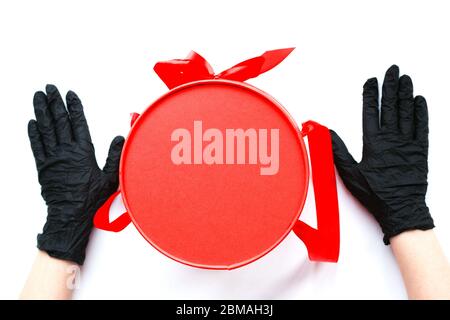 Mani in guanti medici neri e scatola regalo rossa con spazio per la copia. Consegna senza contatto durante l'isolamento durante la pandemia di coronavirus. Foto Stock