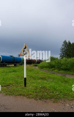 Cartello stradale "attraversamento ferroviario senza barriera" sullo sfondo di un treno con serbatoi di petrolio e orientamento verticale di una strada di campagna Foto Stock