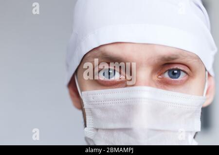 Medico stanco giovane uomo in maschera durante il coronavirus guarda la macchina fotografica, primo piano Foto Stock