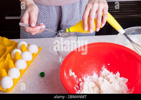 Le mani femminili tengono il cucchiaio e le bottiglie di succo di limone pronto all'uso e lo versano nell'impasto del formaggio casolare e della farina in una ciotola rossa Foto Stock