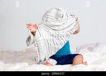 Piccolo ragazzo caucasico che gioca sotto una coperta a righe su lenzuola bianche con piumone Foto Stock