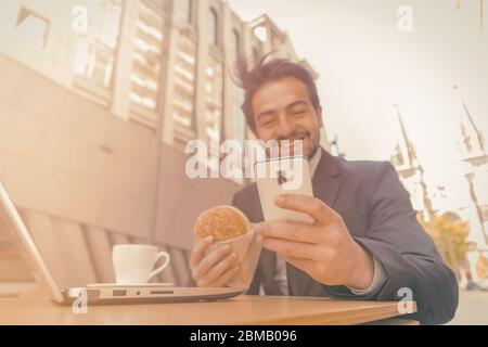 L'uomo d'affari mangia l'hamburger usando il telefono sulla strada. Uomo sorridente in tuta mangiare in fast food caffetteria sulla strada di una moderna città europea. Tonato Foto Stock