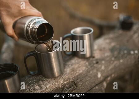 La mano maschile sta versando una bevanda calda da un thermos in coppe da campeggio di metallo in piedi su un albero caduto Foto Stock