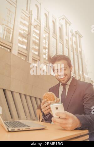 Uomo d'affari che usa il telefono che mangia sandwich o hamburger all'aperto. Happy man in tuta si trova nella caffetteria fast food sulla strada della città. Immagine a toni. Verticale Foto Stock