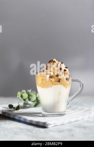 Dalgona in una tazza di vetro trasparente decorata con gocce di cioccolato, bevanda coreana di tendenza fatta con latte e schiuma di caffè su sfondo grigio Foto Stock