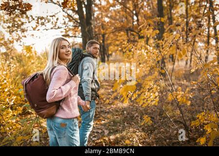 Sorridente coppia di turisti che camminano lungo un sentiero forestale tenendo le mani e guardando indietro la macchina fotografica Foto Stock