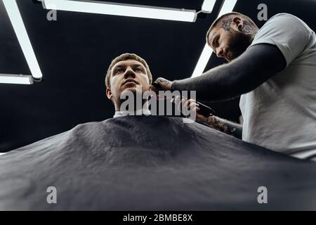 Taglio di capelli maschile con rasoio elettrico. Tattooed Barber fa un taglio di capelli per il cliente al barbiere usando il hairclipper. Uomo parrucchiere con elettrico Foto Stock