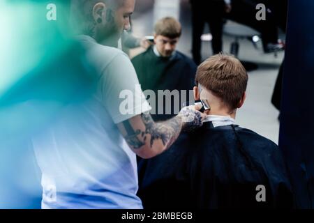 Taglio di capelli maschile con rasoio elettrico. Tattooed Barber fa un taglio di capelli per il cliente al barbiere usando il hairclipper. Uomo parrucchiere con elettrico Foto Stock