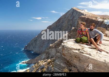Una giovane coppia seduta sul bordo di una scogliera e godendo di una vista costiera a Folegandros, Cicladi, Grecia Foto Stock