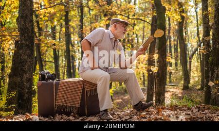 letteratura al di fuori della biblioteca. ottenere conoscenza e informazioni dalla natura. Il vecchio leggeva seduto su una valigia retrò. Viaggio d'autunno. Avere il piacere di leggere di più. Uomo in pensione ha letto il libro in autunno natura. Foto Stock