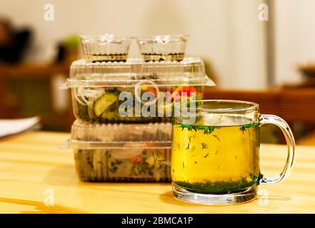 Tazza con brodo caldo ed erbe su un tavolo di legno. Sullo sfondo di contenitori monouso trasparenti con insalata di pomodori e cetrioli. BL Foto Stock