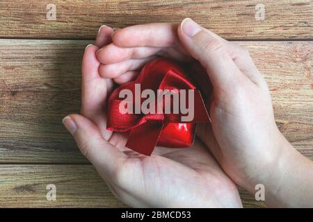 Mani donna che tengono un cuore dono rosso con arco su sfondo di legno. San Valentino, giorno della madre, compleanno, festa, dono Foto Stock