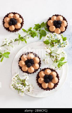 Crostata di cioccolato con ripieno di caramello salato e bacche nere fresche. Fiori di ciliegio su sfondo. Spazio di copia. Foto Stock