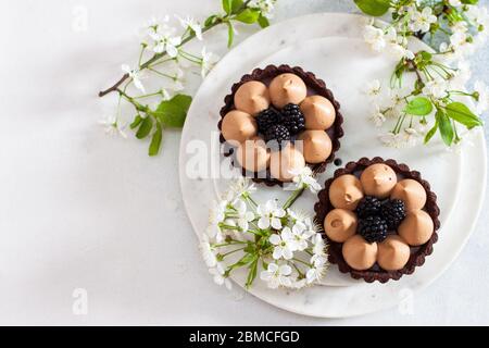 Crostata di cioccolato con ripieno di caramello salato e bacche nere fresche. Fiori di ciliegio su sfondo. Spazio di copia. Foto Stock