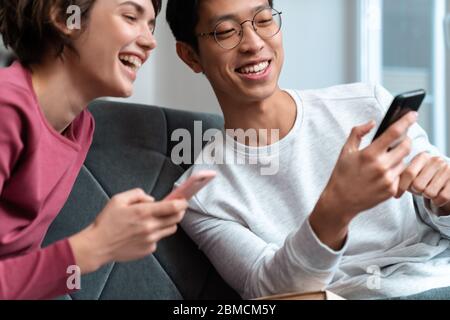 Foto di una coppia multinazionale allegra che ride e usa gli smartphone mentre si siede sul divano a casa Foto Stock
