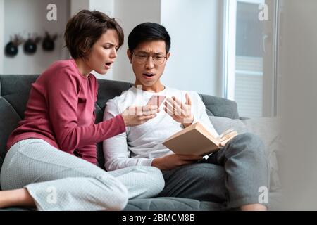 Foto di una coppia multinazionale scioccata che legge il libro e utilizza lo smartphone mentre si siede sul divano a casa Foto Stock