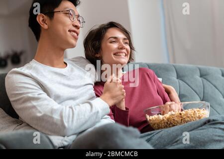 Foto di una coppia multinazionale allegra che mangia popcorn e guarda il film mentre si siede sul divano a casa Foto Stock