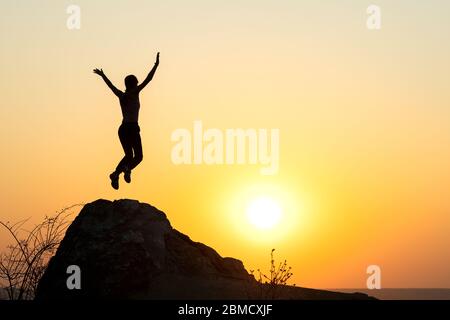 Silhouette di donna escursionista che salta da solo su roccia vuota al tramonto in montagna. Turista femminile che alza le mani in piedi sulla scogliera nella natura serale. Foto Stock