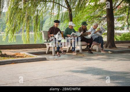 Hanoi Vietnam ottobre 25 2013; quattro uomini vietnamiti anziani seduti insieme sul panca del parco in ombra sotto l'albero di salice dal lago tutti la lettura della loro newspaper Foto Stock