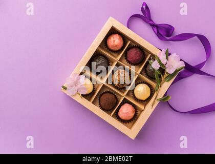 Set di diversi cioccolatini in una scatola di carta con un nastro viola satinato su uno sfondo luminoso. Concetto di vacanza. Foto Stock