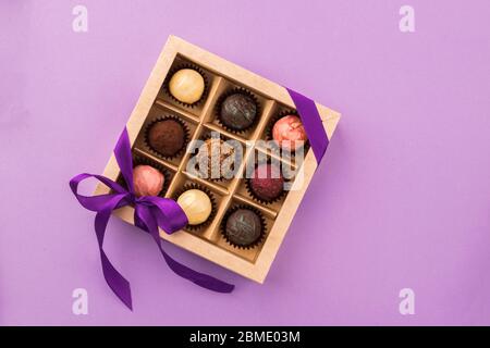 Set di diversi cioccolatini in una scatola di carta con un nastro viola satinato su uno sfondo luminoso. Concetto di vacanza. Foto Stock