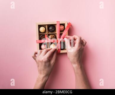 Una donna si stanca un arco e apre una scatola di cioccolatini. Set di diversi cioccolatini in una scatola di carta a mani femminili. Foto Stock