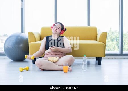 Donna asiatica sovrappeso utilizzando tablet e.. Cuffia per ascoltare musica e lei è l'esercizio a casa, la ragazza asiatica godere mangiare cibo e popcorn Foto Stock