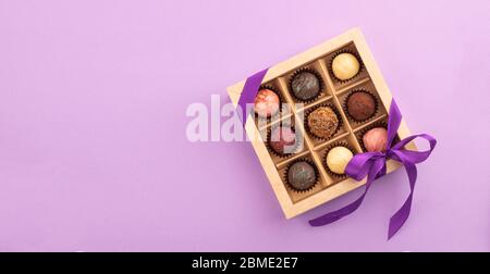 Set di diversi cioccolatini in una scatola di carta con un nastro viola satinato su uno sfondo luminoso. Concetto di vacanza. Foto Stock