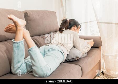 Donna asiatica con occhiali che legge un libro e ascolta la musica sdraiata sul divano con i piedi in su. Giovane ragazza millenaria che legge un vecchio libro a casa. Foto Stock