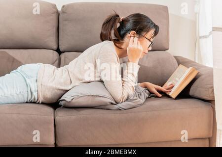 Donna asiatica con occhiali che legge un libro e ascolta la musica sdraiata sul divano con i piedi in su. Giovane ragazza millenaria che legge un vecchio libro a casa. Foto Stock