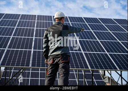 Vista ad angolo basso sul tecnico in uniforme blu scuro con la schiena alla telecamera, puntando verso l'impianto solare. Vista posteriore. Fonte ecologica alternativa di concetto di energia Foto Stock