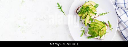 Panino di pane al formaggio cremoso e fette di avocado su un piatto su sfondo chiaro. Cibo vegetariano sano. Vista dall'alto, dall'alto, banner Foto Stock