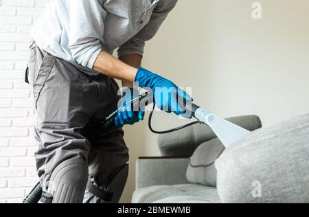 Uomo impiegato di aspirapolvere che rimuove lo sporco dai mobili in un divano piano, pulito sotto vuoto con attrezzature professionali Foto Stock