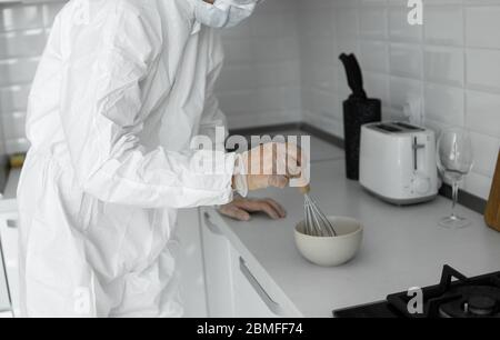 L'uomo in tuta protettiva bianca e in maschera medica cuoce nella cucina bianca a casa durante il coronavirus. Rimani a casa. Goditi la cucina a casa. COVI Foto Stock